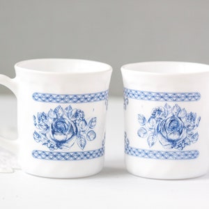 May include: Two white ceramic mugs with a blue floral rose design. Each mug features a handle and a decorative blue band with a geometric pattern. The floral design is centered on the mug's body.