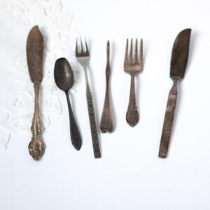 May include: An assortment of antique silverware, including knives, forks, and a spoon, arranged on a white surface. The silverware has a tarnished, vintage appearance, with ornate handle details on some pieces. The arrangement suggests a focus on dining or kitchen decor.