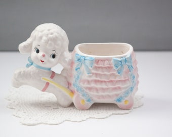 May include: A vintage ceramic poodle planter in white, pink, and blue. The poodle is attached to a pink and blue carriage-like planter with a ruffled design. The planter has a blue bow on each side and is on a white lace doily.
