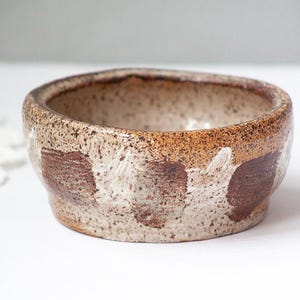 May include: A handmade ceramic bowl with a speckled beige and brown exterior. The bowl features a wide, shallow design with a textured surface and dark brown painted accents. This rustic bowl is suitable for serving or display.