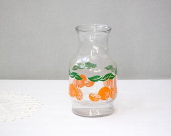 May include: Clear glass pitcher with a wide base and a narrow neck. The pitcher is decorated with orange fruit and green leaf designs. The pitcher is suitable for serving beverages.