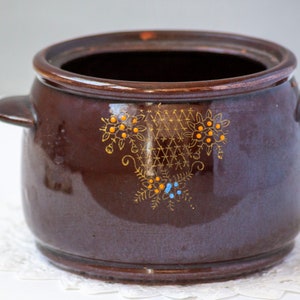 May include: A brown ceramic pot with two side handles and a decorative floral design. The pot has a glossy finish and is adorned with orange and blue floral accents. This vintage kitchenware is ideal for cooking and serving.