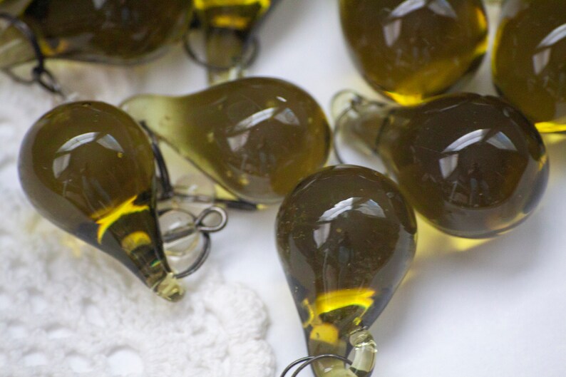 K&ouml;nnte beinhalten: Eine Sammlung olivgr&uuml;ner, tropfenf&ouml;rmiger Glasornamente. Jedes Ornament hat eine kleine Metallschlaufe zum Aufh&auml;ngen. Das Glas hat einen leichten Gelbstich und reflektiert das Licht. Die Ornamente sind wahrscheinlich f&uuml;r dekorative Zwecke.