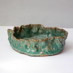 May include: A handcrafted, teal-colored ceramic bowl with a textured, organic design. The rim has a rough, uneven edge with hints of brown. This decorative bowl is suitable for serving or display.