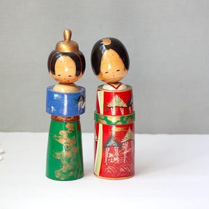 May include: Two vintage Japanese Kokeshi dolls. One doll has a gold-colored head, blue midsection, and green base. The other doll has a black head, red midsection, and green base. Both dolls have painted facial features and are on a white surface.