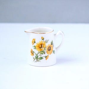 May include: A small, white ceramic creamer with a handle and a gold rim. The creamer is decorated with a floral pattern of yellow sunflowers and green leaves. The creamer is on a white surface.