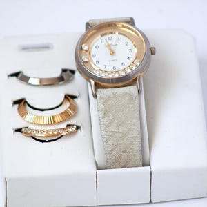 May include: A gold-tone analog wristwatch with a white face, Arabic numerals, and a textured beige strap. The watch bezel is accented with clear crystals. Included are three interchangeable gold-tone metal watch bezels, one with crystals.
