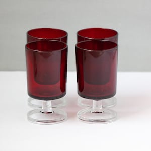 May include: Four ruby red glass candle holders with clear glass stems. The candle holders are cylindrical with a flared base. The glasses are arranged in a row on a white surface.