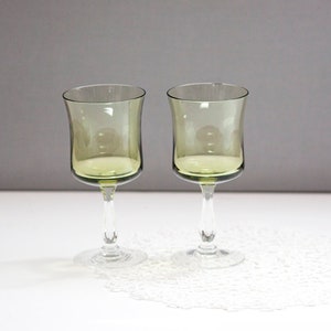 May include: Two green-tinted wine glasses with clear stems and bases. The glasses have a tulip-like shape and sit on a white, lacy doily. The background is a solid gray.