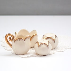 May include: A cream-colored ceramic creamer, sugar bowl, and two salt and pepper shakers. The set has gold trim and sits on a white lace doily. The creamer has a gold handle.