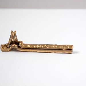 May include: A gold-colored metal nutcracker with a squirrel design. The nutcracker is lying horizontally on a white surface. The squirrel is at the top of the nutcracker, and the handle is textured.