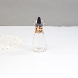 May include: A clear glass bottle with a black stopper and gold-colored detailing around the neck. The bottle has a conical shape and is empty. The background is a plain white surface.
