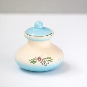 May include: A small, ceramic jar with a lid. The jar is a light cream color with a blue base and lid. A floral design with pink and white flowers and green leaves decorates the front. The jar is likely used for storage.