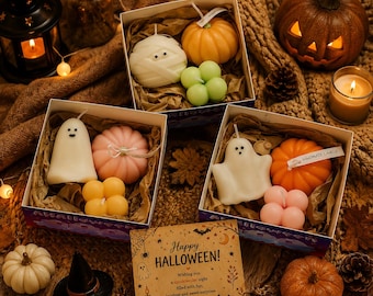 May include: Three square gift boxes containing assorted Halloween-themed candles shaped like ghosts, pumpkins, and small spheres. A square card with the text Happy HALLOWEEN and illustrations of a cat, ghost, and pumpkin sits in the foreground.