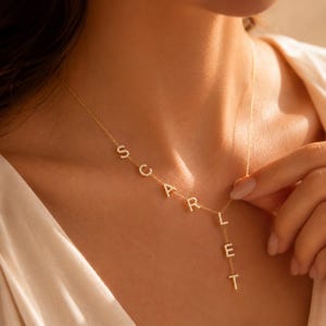 May include: A gold-toned lariat necklace featuring the name SCARLET spelled out in individual, pavé-set crystal letters. The letters S, C, A, R, L are arranged along the chain, while the letters E and T hang vertically from the centre point.