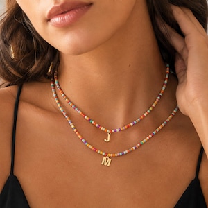 May include: A layered necklace set featuring two strands of small, multicoloured glass beads. The shorter strand includes a gold-toned letter J pendant, while the longer strand features a gold-toned letter M pendant, both centred on the neck.