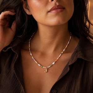 Puede incluir: Un collar de cuentas con un colgante en forma de letra J dorada. El collar está compuesto por pequeñas cuentas de vidrio multicolor intercaladas con pequeñas perlas blancas de forma ovalada y diminutas cuentas espaciadoras metálicas doradas.