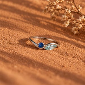 Peut inclure: Une bague argentée dotée d'un anneau incurvé. La bague est ornée d'une petite pierre précieuse ronde bleu foncé et d'une pierre plus grande bleu clair taille marquise, toutes deux placées sur la courbe supérieure de l'anneau en métal poli.
