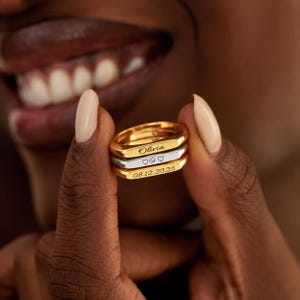 Peut inclure: Un empilement de trois bagues en métal avec des surfaces avant plates et rectangulaires. La bague supérieure est dorée et gravée du prénom Olivia. La bague centrale est argentée et gravée d'un cœur, de la lettre G et d'un cœur. La bague inférieure est dorée et gravée de la date 08.12.2025.