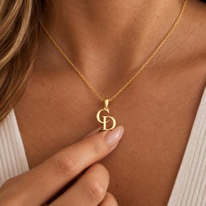 May include: A gold-toned necklace featuring a pendant with the interlocking capital letters C and D. The pendant is suspended from a delicate, thin gold-toned chain that rests against the collarbone, showcasing a polished metallic finish throughout.