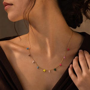 Op de afbeelding: Een goudkleurige ketting met kleine, veelkleurige letterbedels die langs de ketting zijn verdeeld. De letters hebben verschillende felle kleuren, waaronder blauw, roze, wit, geel en groen, hangend aan een fijne metalen schakelketting.