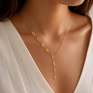May include: A gold-toned necklace featuring a delicate chain with the name SCARLETT spelled out in individual capital letters. The letters S, C, A, R, L are arranged diagonally along the chain, while the letters E, T, T are suspended vertically.
