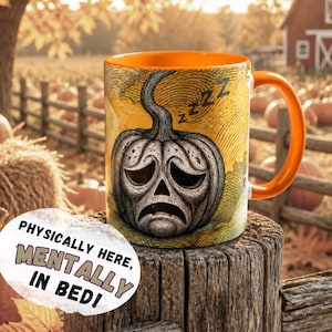 Könnte beinhalten: Orangefarbene Keramiktasse mit einem Design eines traurigen Kürbisgesichts. Die Tasse hat einen orangefarbenen Henkel und Innenraum. Der Text auf der Tasse lautet: „Physisch hier, mental im Bett!“ Der Hintergrund zeigt einen Kürbisacker und einen Holzzaun.