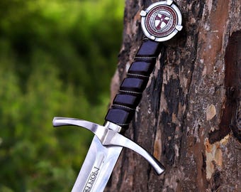 May include: A medieval sword with a silver blade and a black leather-wrapped handle. The hilt features a crossguard and a decorative pommel with a red and white emblem. The blade has the inscription "NON NOBIS, DO".
