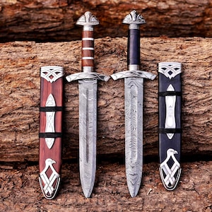 May include: Two daggers with patterned Damascus steel blades and silver-toned metal pommels and guards. One dagger has a brown wood handle and matching scabbard, while the other has a black handle and scabbard. Both scabbards feature silver bird motifs.
