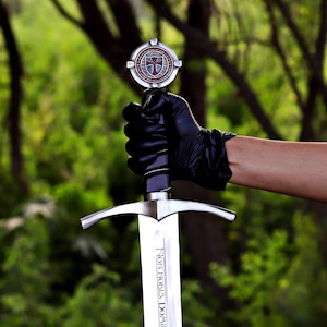 Puede incluir: Espada plateada con empu&ntilde;adura decorativa y emblema de escudo. La hoja de la espada tiene la inscripci&oacute;n "Non Nobis, Domine". La espada es sostenida por una mano enguantada sobre un fondo verde borroso.