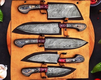 May include: A set of five kitchen knives with Damascus steel blades and dark wood handles with red accents, each secured in a tan leather roll-up case. The knives vary in size and shape, featuring distinct wavy patterns on the metal blades.