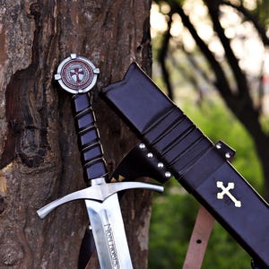 Puede incluir: Una espada medieval con una hoja plateada y un mango negro, apoyada en un &aacute;rbol. La espada tiene una empu&ntilde;adura en forma de cruz y una vaina con una cruz dorada. La hoja lleva la inscripci&oacute;n "NON NOBIS".