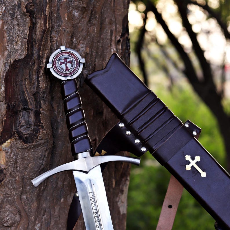 Puede incluir: Una espada medieval con una hoja plateada y un mango negro, apoyada en un &aacute;rbol. La espada tiene una empu&ntilde;adura en forma de cruz y una vaina con una cruz dorada. La hoja lleva la inscripci&oacute;n "NON NOBIS".