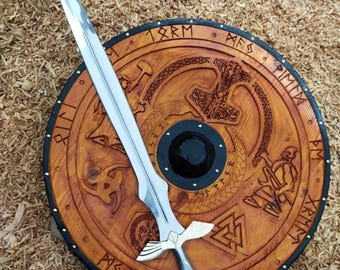 May include: A silver-toned sword with a metallic hilt resting on a circular wooden shield. The shield features carved Norse-inspired symbols, runes along the outer edge, and a central black metal boss. The sword has a pointed blade and crossguard.