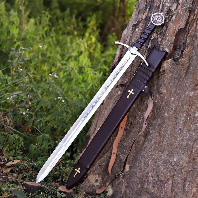 Puede incluir: Una espada plateada con empu&ntilde;adura negra y guarda en forma de cruz descansa contra un &aacute;rbol. La hoja de la espada tiene texto grabado. Una funda de cuero marr&oacute;n con cruces doradas y una correa est&aacute; junto a la espada.