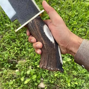Op de afbeelding: Een mes met een vaststaand lemmet, voorzien van een getextureerd, donkergrijs metalen blad met een gepolijste snede en een messing stootplaat. Het handvat is gemaakt van natuurlijk bruin hertengewei met een witte kern en een ruw oppervlak.