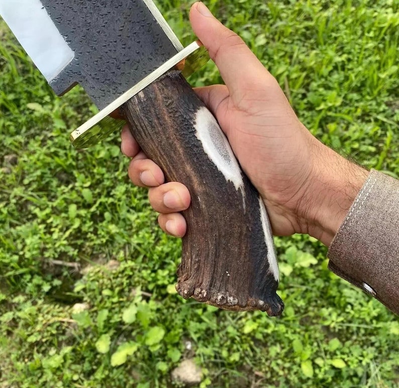 Op de afbeelding: Een mes met een vaststaand lemmet, voorzien van een getextureerd, donkergrijs metalen blad met een gepolijste snede en een messing stootplaat. Het handvat is gemaakt van natuurlijk bruin hertengewei met een witte kern en een ruw oppervlak.
