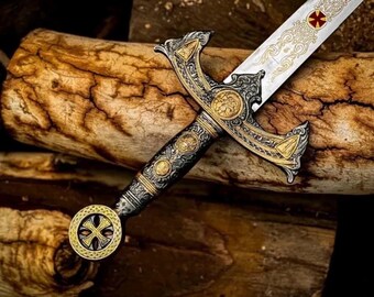 May include: Ornate sword with a silver blade and gold accents. The hilt features intricate designs and a circular pommel. The blade has a decorative pattern and a red cross emblem. The sword rests on a textured wooden surface.