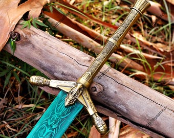 Puede incluir: Una espada decorativa con una empuñadura de metal dorado con intrincados patrones grabados y una guarda. La hoja es de un color verde azulado vibrante y muestra diseños grabados detallados a lo largo, apoyada sobre un tronco de madera.