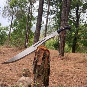 Puede incluir: Un machete grande de metal plateado con una hoja larga y curva que presenta un lomo dentado y un canal central. El mango es negro con una empuñadura texturizada y remaches plateados, que se extiende desde un pomo curvo.