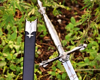Puede incluir: Una daga de metal plateado con un pomo puntiagudo, un mango estriado y una guarda con extensiones angulares afiladas. La hoja muestra un recorte decorativo central. Junto a ella se encuentra una vaina negra con una boquilla de metal plateado.