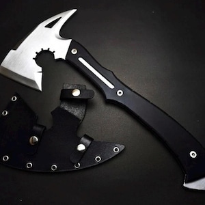 May include: A silver-colored metal hatchet with a curved blade and a cutout design near the head. The handle is black with a silver stripe and two rivets. A black leather sheath with silver metal eyelets and snap buttons is positioned below.
