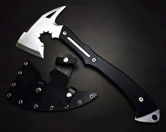 May include: A silver-colored metal hatchet with a curved blade and a cutout design near the head. The handle is black with a silver stripe and two rivets. A black leather sheath with silver metal eyelets and snap buttons is positioned below.