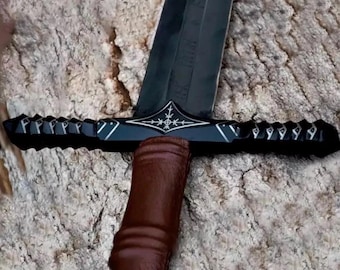 Puede incluir: Una espada negra con una hoja oscura y un mango envuelto en cuero marrón. La guarda cruzada presenta un diseño decorativo con una incrustación plateada. La espada está sobre una superficie texturizada de color marrón claro.
