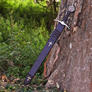 Puede incluir: Una espada medieval con una vaina negra, apoyada contra un &aacute;rbol. La espada presenta una guarda cruzada plateada y una empu&ntilde;adura decorativa con un emblema circular. La vaina est&aacute; adornada con cruces doradas y una correa de cuero.