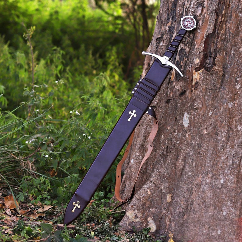Puede incluir: Una espada medieval con una vaina negra, apoyada contra un &aacute;rbol. La espada presenta una guarda cruzada plateada y una empu&ntilde;adura decorativa con un emblema circular. La vaina est&aacute; adornada con cruces doradas y una correa de cuero.