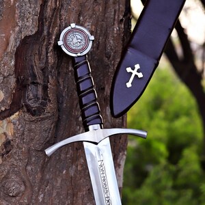 Puede incluir: Una espada medieval con una hoja plateada y un mango negro. La espada tiene una guarda en forma de cruz y un pomo decorativo. La vaina es negra con una cruz blanca. La hoja dice "Non Nobis Domine".