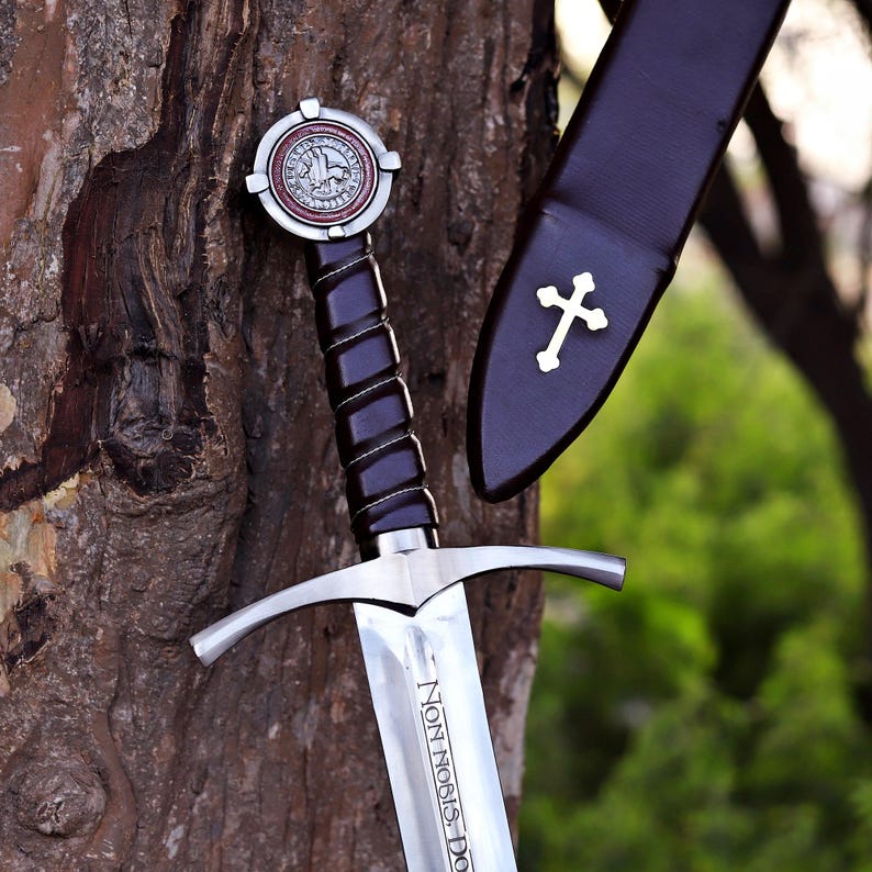 Puede incluir: Una espada medieval con una hoja plateada y un mango negro. La espada tiene una guarda en forma de cruz y un pomo decorativo. La vaina es negra con una cruz blanca. La hoja dice "Non Nobis Domine".