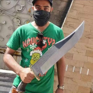 Könnte beinhalten: Ein großes Messer mit feststehender Klinge, das eine texturierte, dunkelmetallische Oberfläche im oberen Bereich und eine glatte, polierte Schneide aufweist. Der Griff besteht aus dunklem, texturiertem Geweih mit messingfarbenem Parier.