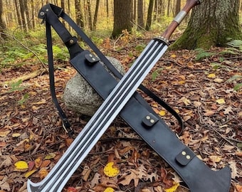 May include: A double-edged sword with a wide, flared tip and three distinct longitudinal grooves along the blade. The handle is crafted from dark wood with a metallic pommel, accompanied by a black leather scabbard featuring adjustable straps.